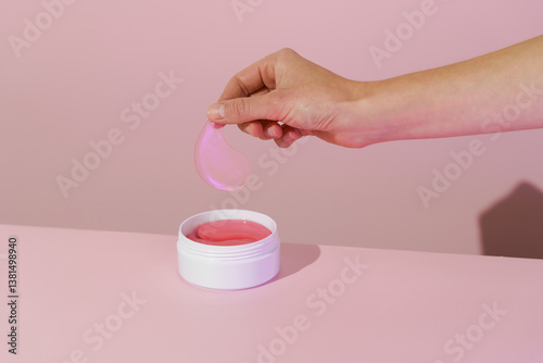 Foto Female hand holding pink hydrogel eye patch over white jar with patches on pink isolated background