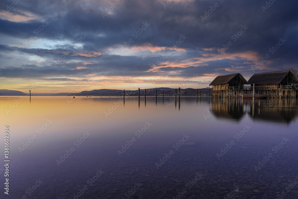 Obraz premium Tranquil sunrise over calm water with wooden cabins on the shore near a peaceful lake in early morning light