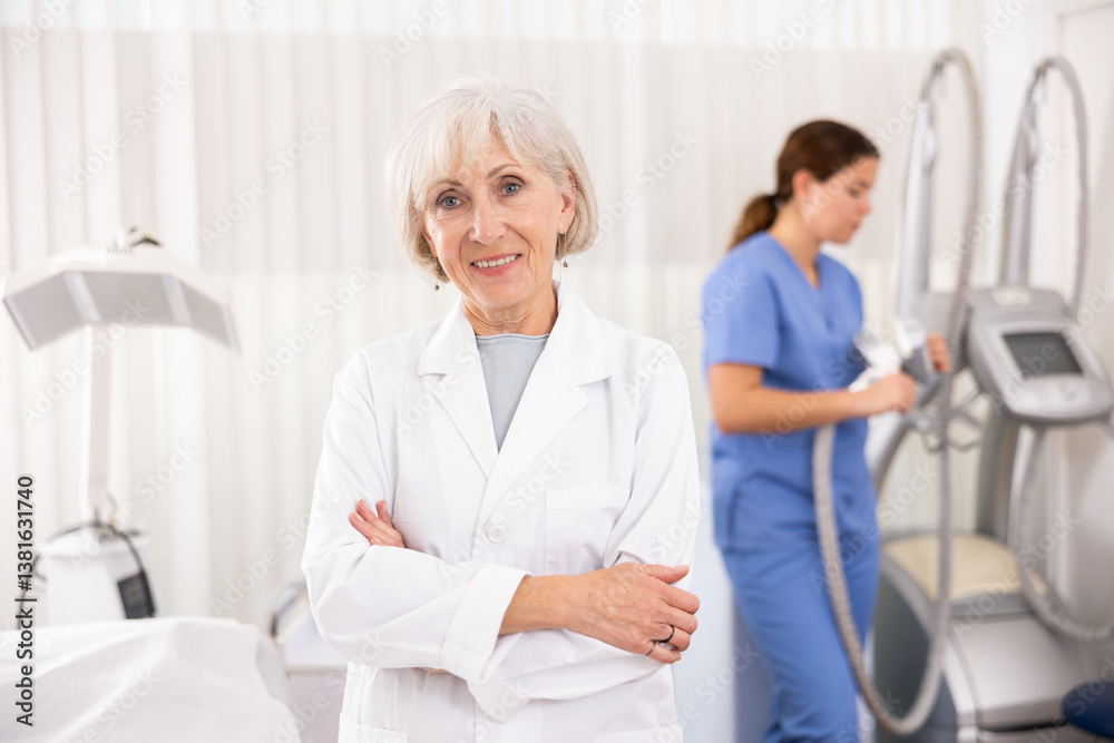 Obraz premium Elderly woman cosmetologist looking at camera while her assistant nurse adjusting vacuum massage machine in clinic of aesthetic medicine