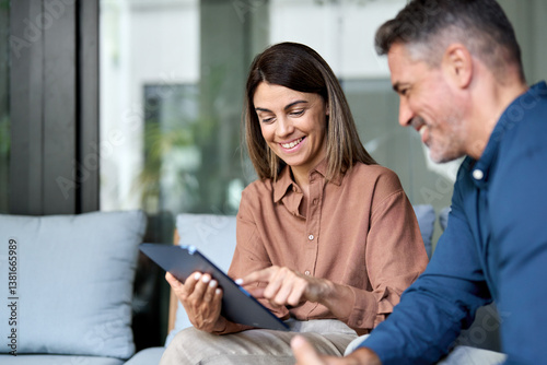 Two happy professional business colleagues working using digital tablet and talking sitting in office on sofa. Smiling mature executives having conversation, manager and client at corporate meeting.