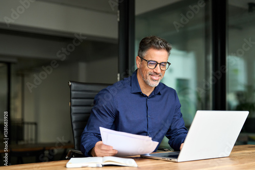 Smiling man executive manager or business owner of middle age reading paper working on computer. Happy busy old professional businessman entrepreneur using laptop holding corporate document in office.