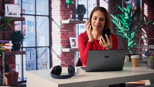 Mature adult smiling and waving during a video call at home desk, showcasing modern technology and online communication in a digital and wireless lifestyle. Web conference on phone. Camera B.
