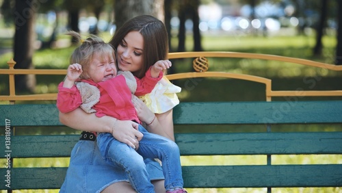 Caring mother soothing distressed child, seated on park bench under bright sunlight