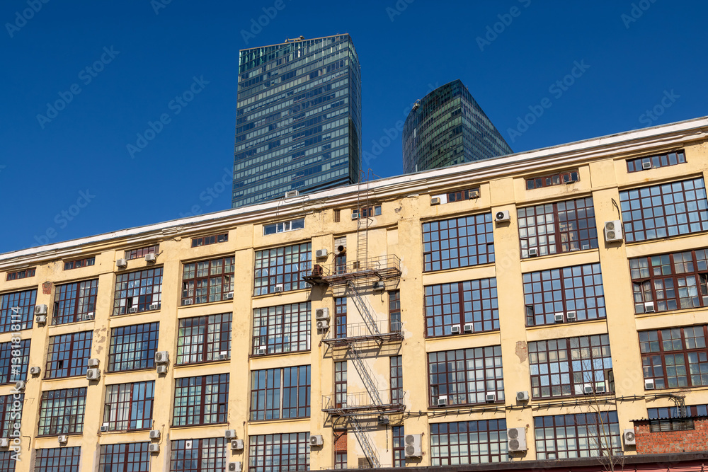 View of the wall of an industrial building, behind which skyscrapers rise. Buildings against the blue sky.