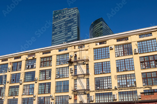 View of the wall of an industrial building, behind which skyscrapers rise. Buildings against the blue sky.