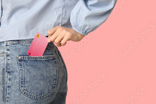 Young woman with credit card in jeans pocket on pink background, closeup