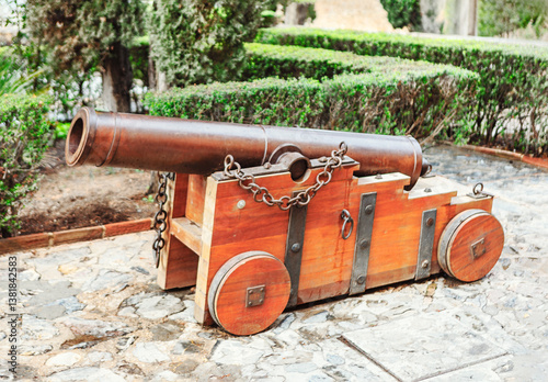 Obraz na płótnie Historic iron cannon on a sturdy wooden carriage, maritime and fortress defense. Set on a stone pathway with lush greenery, it adds an authentic touch to historical and military-themed collections w salonie