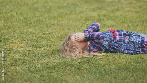 Upset child lying on verdant lawn, hands shielding face, conveying emotional isolation and overwhelming sadness