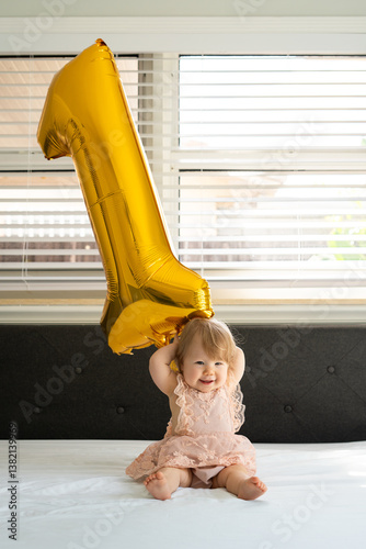 Joyful baby girl celebrates her first birthday while sitting on a white bed, beside a large golden number one balloon. Baby's first birthday. Baby to toddler. Baby's first year. 