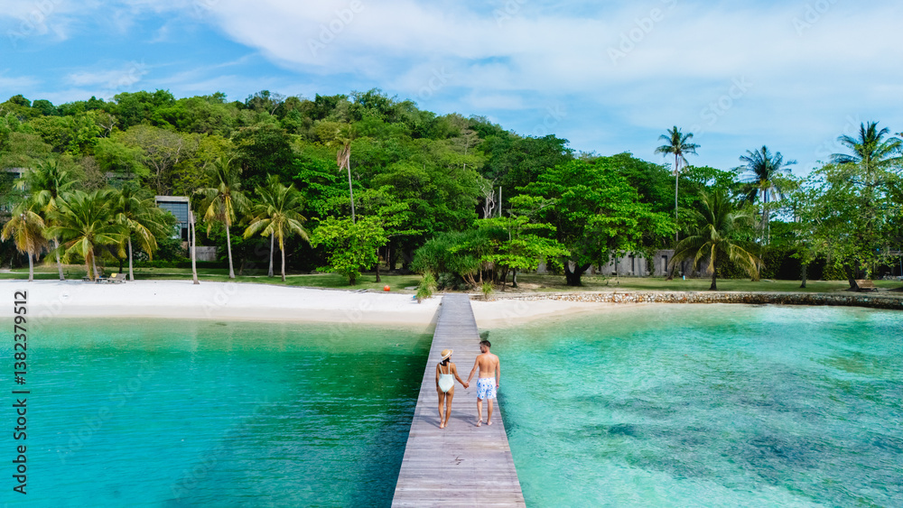 A romantic couple walks hand in hand along a wooden pier, surrounded by crystal-clear waters and lush greenery. The serene atmosphere captures the essence of paradise on Koh Kham Island Thailand A romantic couple walks hand in hand along a wooden pier, surrounded by crystal-clear waters and lush greenery. The serene atmosphere captures the essence of paradise on Koh Kham Island Thailand