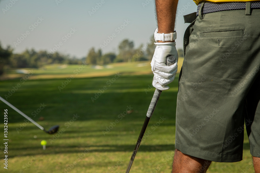 Obraz premium Closeup of an man wearing green khaki shorts and a golf glove holding a golf club with the golf course in the background. Another golfer is in the background