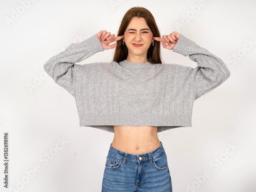 Happy Caucasian young girl wearing grey sweater  ignores loud music and plugs ears with fingers asks to turn off sound
