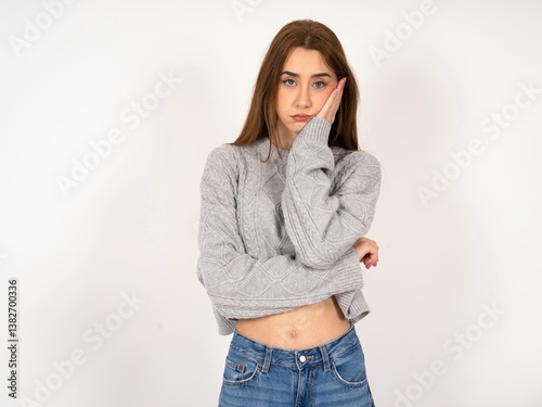 Sad lonely Caucasian young girl wearing grey sweater touches cheek with hand bites lower lip and gazes with displeasure. Bad emotions