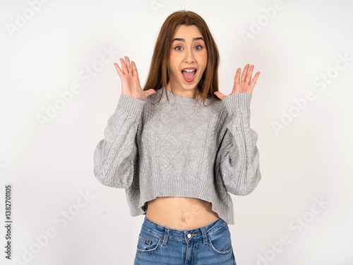 Surprised happy Caucasian young girl wearing grey sweater , glad to see big discounts on clothes, expresses shock, keeps hands near head, jaw dropped.