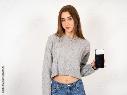 Caucasian young girl wearing grey sweater holds new mobile phone and looks mysterious aside shows blank display of modern cellular