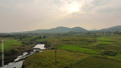 beautiful view of a serene river surrounded by lush green fields, distant hills, and a glowing sunset creating a tranquil and picturesque atmosphere. small dam on river to stop & slow down water flow