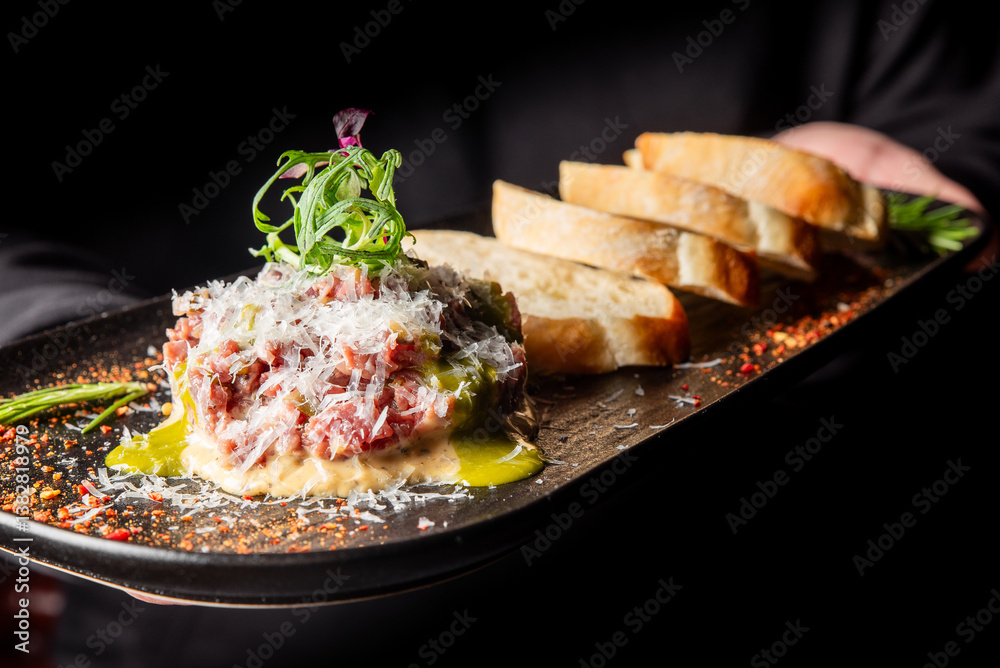 A server presents a gourmet beef tartare topped with microgreens and grated cheese, accompanied by slices of toasted bread on a stylish black platter, garnished with herbs.