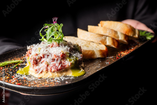 Obraz na płótnie A server presents a gourmet beef tartare topped with microgreens and grated cheese, accompanied by slices of toasted bread on a stylish black platter, garnished with herbs. w salonie