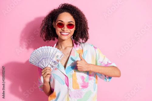 Confident young woman in colorful shirt holding dollar bills giving thumbs up against pink background