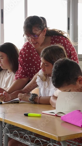 Vertical HD video of young female teacher assisting elementary student girl during lesson in classroom at primary school. Education concept.