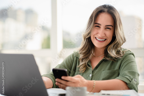Cheerful Latin businesswoman in her 30s wearing green button-up shirt using smartphone while working on laptop in bright modern office with urban skyline view through large windows