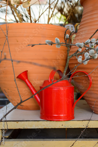bright red watering can stands on the table near clay vases and willow branches