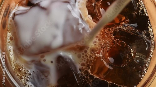 Milk pours into iced black coffee in macro view, creating creamy swirls, bubbles, and textures among glossy ice cubes in a clear glass.