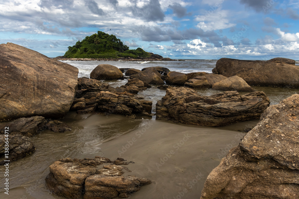 Rocky tropical shore with visible barnacles (Cirripedia) on the stones at low tide. In the background, a lush green island adds depth to this coastal landscape scene.