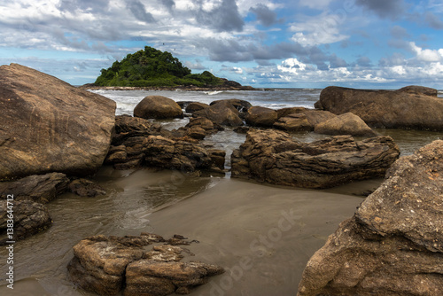 Obraz na płótnie Rocky tropical shore with visible barnacles (Cirripedia) on the stones at low tide. In the background, a lush green island adds depth to this coastal landscape scene. w salonie