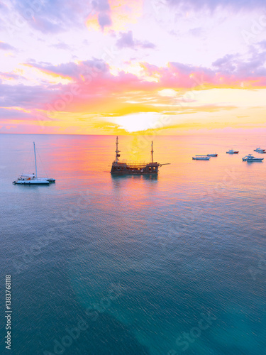 Aerial view of colorful sunrise over ocean in Punta Cana. Boats anchored in calm waters, surrounded by breathtaking scenery. Sky is painted with stunning hues, and gentle waves reflect morning light