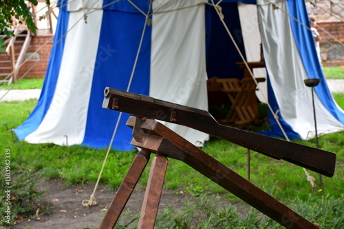 A close up on an old medieval rifle or bombard with a metal barrel and wooden handle laying on a wooden frame, next to a cloth tent and some medieval knight jackets seen in summer in Poland