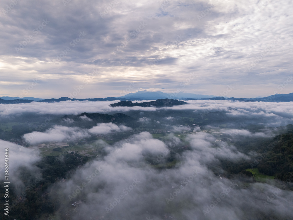 An wide Aerial view of a mountainous landscape with green fields and a village in the foreground. The mountain dominates the background, partially covered by clouds. Tana Toraja - Indonesia An wide Aerial view of a mountainous landscape with green fields and a village in the foreground. The mountain dominates the background, partially covered by clouds. Tana Toraja - Indonesia