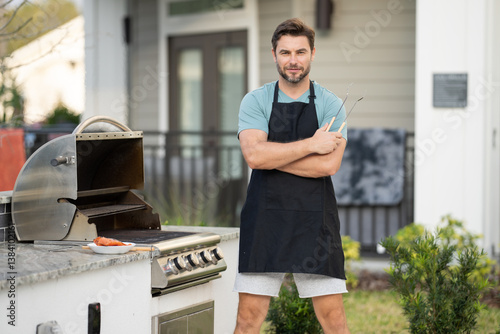 male chef in an apron stands by the grill outside and looks at camera. man is cooking on a grill. chef man at barbecue and grill . chef at barbecue and grill. Man grilling food on barbecue in backyard