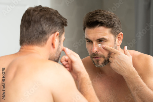 Handsome Man Applying Face Cream in the Bathroom. Closeup beauty portrait of satisfied young man after facial cream. Attractive brunet guy touching skin. Male coscmetic and beauty concept
