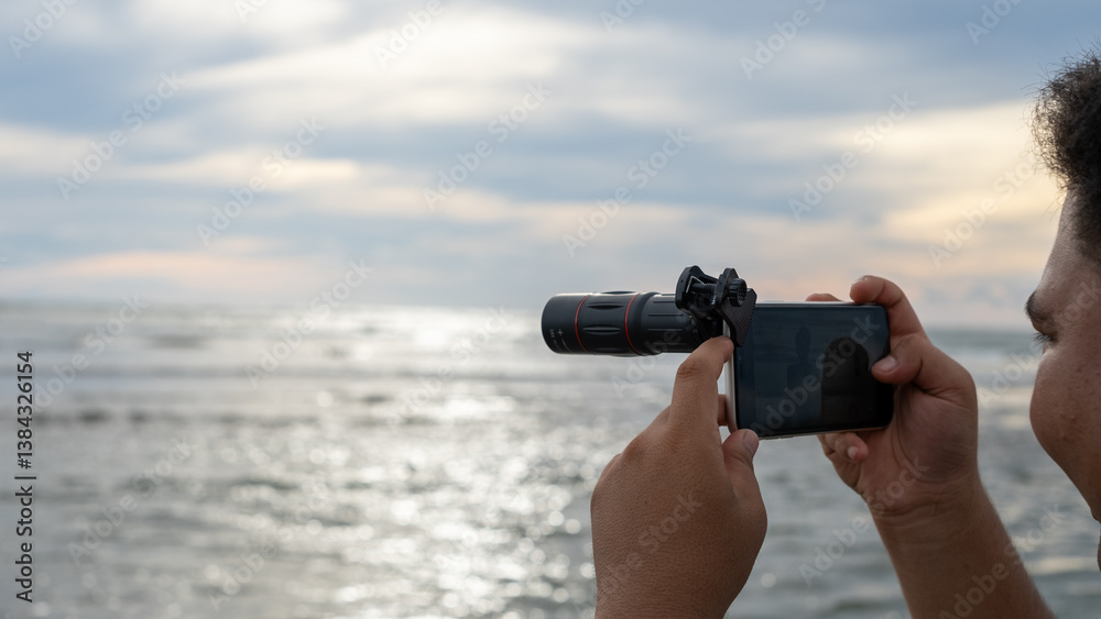A person using a smartphone with a telephoto lens attachment to capture the ocean view during sunset. A person using a smartphone with a telephoto lens attachment to capture the ocean view during sunset.
