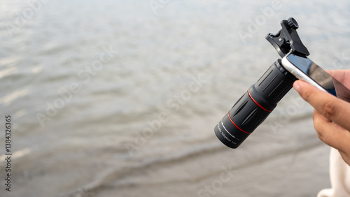 A close-up of a smartphone with a telephoto lens attachment, held over calm seawater. The compact clip-on lens enhances mobile photography, showcasing innovation in portable optics.