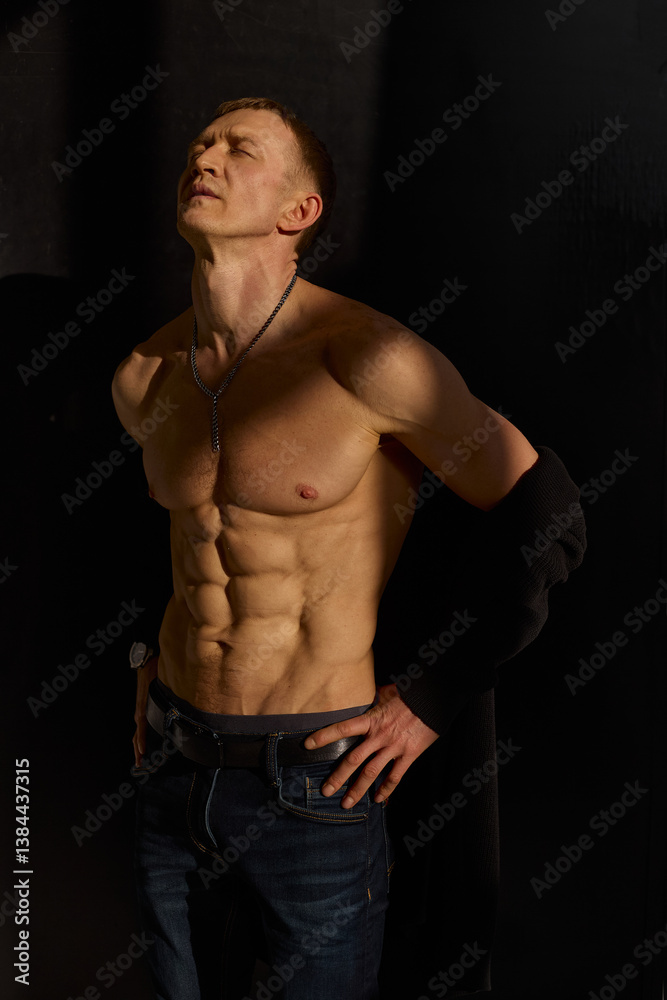Fototapeta premium Slim muscular male model on black background. Shirtless fitness guy in black sweatpants posing in studio. Man with six pack abs in sunset light. Brutal photo
