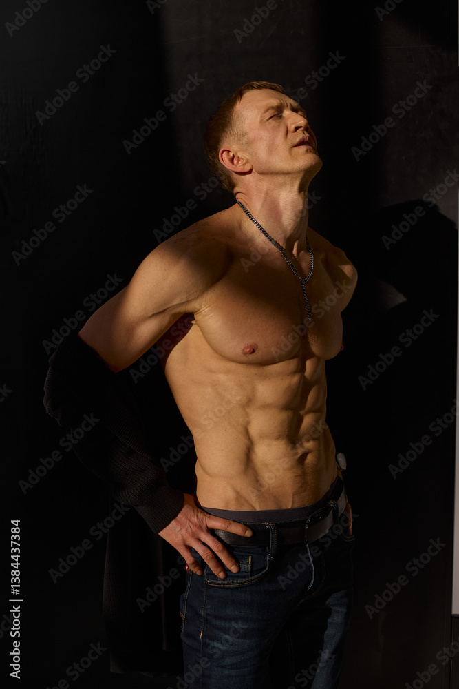 Fototapeta premium Slim muscular male model on black background. Shirtless fitness guy in black sweatpants posing in studio. Man with six pack abs in sunset light. Brutal photo