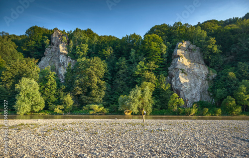 Canvas Print Rear view of a bearded traveler with a backpack standing on a rocky riverbank, admiring the scenic view of lush green mountains and towering rock formations