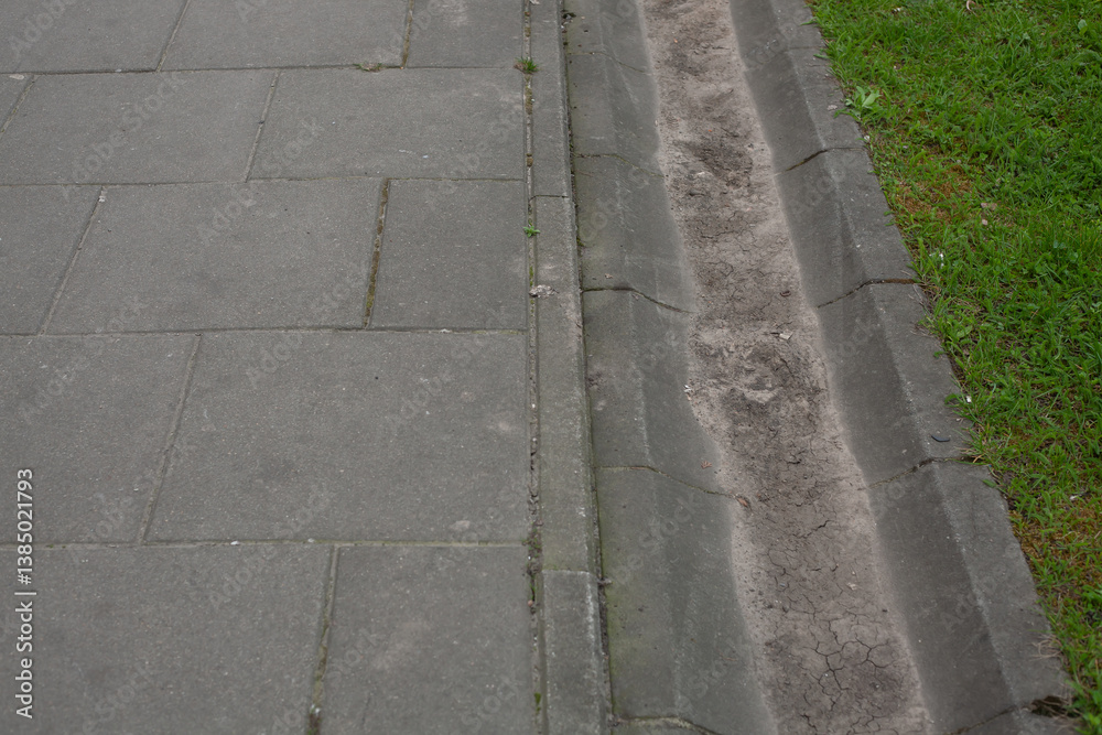 Urban sidewalk with concrete slabs and shallow drainage channel along the lawn. Typical pedestrian infrastructure and water runoff system in a city, top-down view Urban sidewalk with concrete slabs and shallow drainage channel along the lawn. Typical pedestrian infrastructure and water runoff system in a city, top-down view