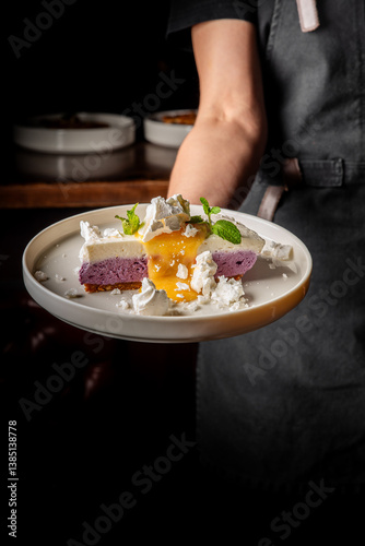 A person holding a plate with a slice of layered dessert topped with whipped cream, mint leaves, and a yellow sauce. The dessert has a purple layer and a white layer, with crumbs scattered around.