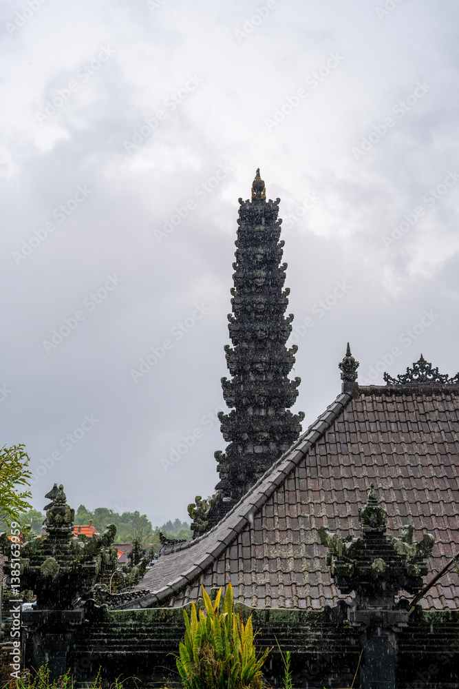 A majestic Balinese temple tower rises amidst a rainy sky, showcasing intricate architecture and cultural heritage. The lush greenery complements the serene atmosphere.