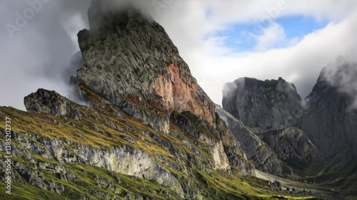 A dramatic view of a towering mountain peak surrounded by mist and clouds, with rugged cliffs and lush green meadows, showcasing the raw beauty of alpine landscapes and natural wilderness.