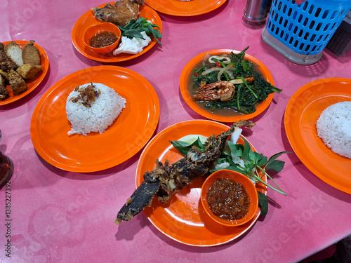 A table at a food stall with pecel lele, sambal, and fresh vegetables on a plate. Indonesian street food.