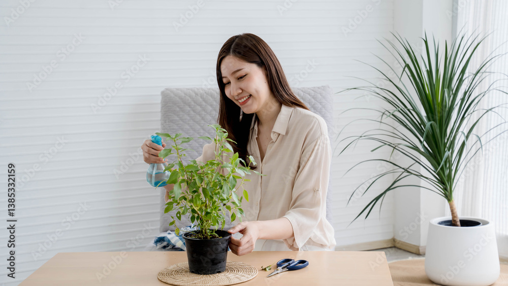 Smiling young Asian woman watering a potted plant with spray bottle while sitting at a wooden table indoors, enjoying peaceful home gardening activity. Smiling young Asian woman watering a potted plant with spray bottle while sitting at a wooden table indoors, enjoying peaceful home gardening activity.