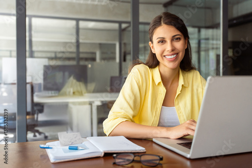 Portrait at camera smiling latin female marketing manager, professional it specialist working at laptop computer sitting at desk in modern office. Cheerful young woman employee using pc for business 