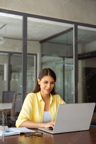 Focused smiling latin female marketing manager, professional ai specialist working at laptop computer sitting at desk in modern office. Young woman student using pc for online business studying course