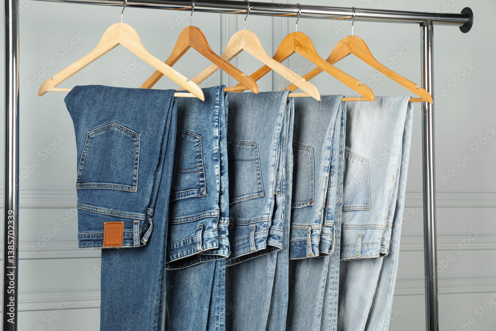Many stylish jeans hanging from rack near white wall