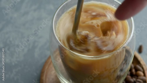 Iced coffee with milk is stirred in a clear glass, creating swirls and smooth texture, viewed in close-up from above.