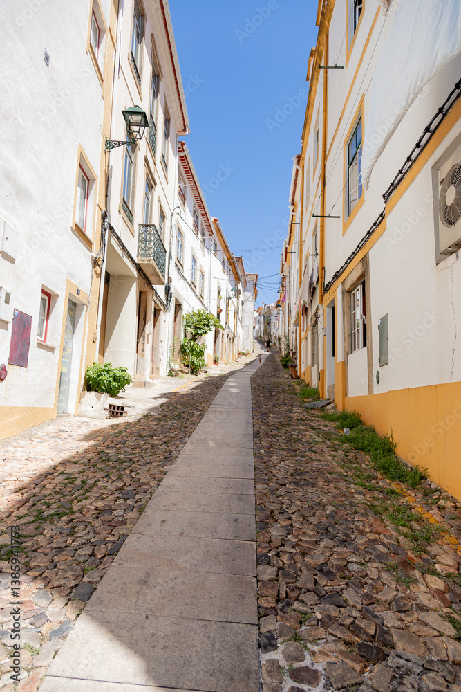 Obraz premium On a sunny afternoon in Castelo de Vide, Portugal, a quiet cobblestone street winds through the historic village of Alentejo, revealing traditional whitewashed houses and yellow details.